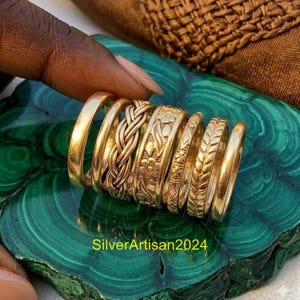 May include: A set of seven gold-toned rings featuring various designs including a smooth band, a braided pattern, a floral motif, a geometric pattern, and a leaf-like texture. The text SilverArtisan2024 is visible at the bottom of the image.