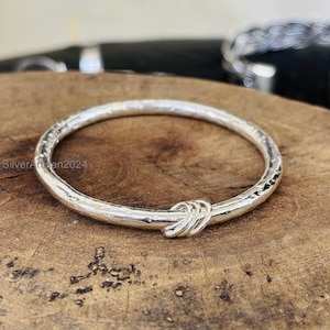 May include: A silver bangle bracelet with a textured surface and a knot design. The bracelet is made of a shiny, metallic material and is displayed on a wooden surface. The text "SilverArtisan2024" is visible.