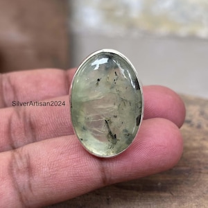 May include: Oval-shaped prehnite gemstone ring with a silver band. The light green stone has dark inclusions and is set in a polished silver bezel. The ring is displayed on a person's fingers.