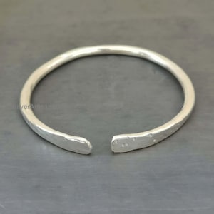 Peut inclure: Un bracelet manchette ouvert de couleur argentée. Le bracelet présente une texture martelée et un design simple et minimaliste. Les extrémités du bracelet sont plates et légèrement arrondies. Le bracelet est posé sur une surface grise.
