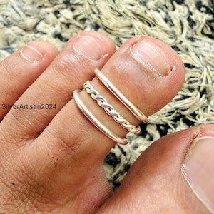Op de afbeelding: Een set van drie zilverkleurige ringen gedragen om een teen. De set bevat twee gladde, effen banden en een centrale gedraaide ring in touwstijl. De ringen zijn op de teen gestapeld en hebben een metalen afwerking en een eenvoudig ontwerp.