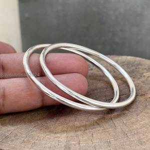 May include: Two silver bangles with a smooth, rounded design. The bangles are displayed on a wooden surface. The image is well-lit, highlighting the reflective quality of the metal. The text "SilverArtisa 2024" is visible.