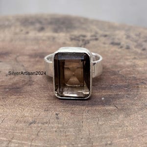 Puede incluir: Un anillo de plata con una gran gema rectangular de color marrón ahumado. El anillo tiene una banda gruesa y un diseño sencillo. La gema está engastada en un bisel. El anillo se muestra sobre una superficie de madera.