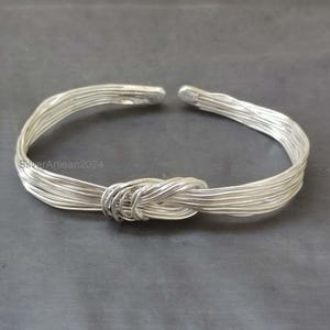 Può includere: Un bracciale a polsino color argento con un design a nodo. Il bracciale è realizzato con più fili di filo metallico, intrecciati per formare un nodo al centro. Le estremità del bracciale sono aperte.