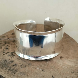 May include: A wide, polished silver cuff bracelet. The bracelet has a smooth, reflective surface and a simple, open design. The cuff is approximately 2 inches wide.