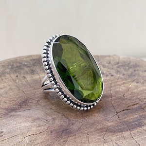 Puede incluir: Anillo ovalado con una piedra preciosa verde oliva facetada, engastada en una banda de plata. La banda tiene un borde de cuentas y un diseño de caña dividida. El anillo se exhibe sobre una superficie de madera. La piedra mide unos 3 cm.