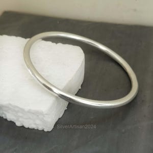 Puede incluir: Un brazalete simple de plata. El brazalete es liso y brillante, con una forma redondeada.