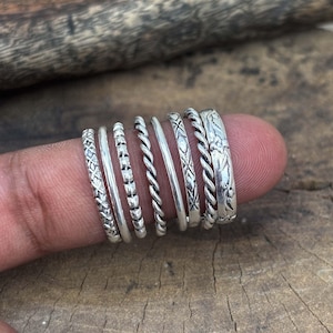 Può includere: Una collezione di anelli d'argento con vari design, tra cui motivi strutturati, corde intrecciate e dettagli floreali. Gli anelli sono esposti su un dito, mostrando i loro diversi stili e trame. Gli anelli sono in argento.