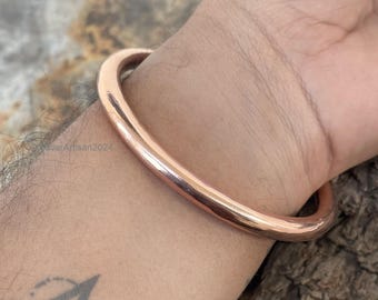 May include: A simple copper bangle bracelet, worn on a person's wrist. The bracelet is smooth and shiny, with a rounded shape.