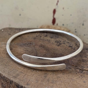May include: A silver-colored, open-ended cuff bracelet. The bracelet has a smooth, rounded band that tapers into two flat, rectangular ends. The bracelet is shown on a wooden surface. The text "SilverArtisan2024" is visible.