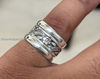 Könnte beinhalten: Nahaufnahme eines silbernen Rings mit floralem Design. Der Ring hat drei Bänder, wobei das mittlere Band ein detailliertes Blumen- und Blättermuster aufweist. Der Ring wird an einem Finger getragen und zeigt seine aufwendige Handwerkskunst.
