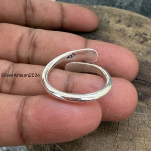 May include: A silver-colored open-ended ring with a smooth, polished finish. The inner band features a clear stamp reading 925, indicating sterling silver composition. The ring has a simple, curved design that overlaps slightly at the ends.