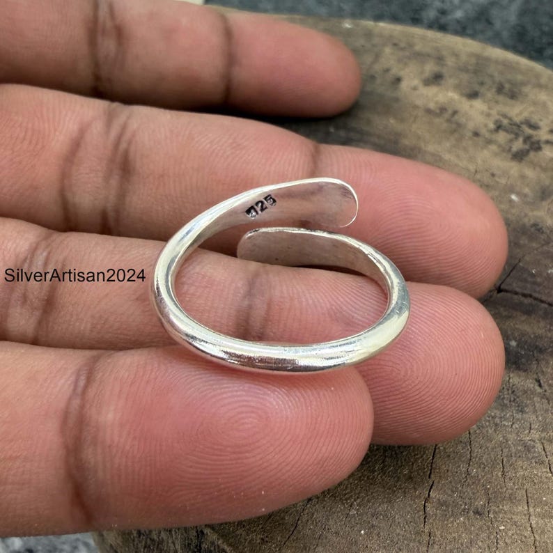 May include: A silver-colored open-ended ring with a smooth, polished finish. The inner band features a clear stamp reading 925, indicating sterling silver composition. The ring has a simple, curved design that overlaps slightly at the ends.