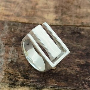 May include: A silver ring with a geometric design. The ring features a rectangular center piece framed by a square shape with open sides. The ring is made of a shiny, metallic material and is set against a wooden background.