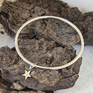 May include: A silver bangle bracelet with a small star charm hanging from it. The bracelet is on a wooden surface.