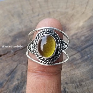 Può includere: Un anello in argento con una pietra ovale traslucida color ambra incastonata in un bordo metallico a corda intrecciata. La fascia è composta da due fili d'argento sottili e lisci che si separano e si ricongiungono ai lati con decorazioni.