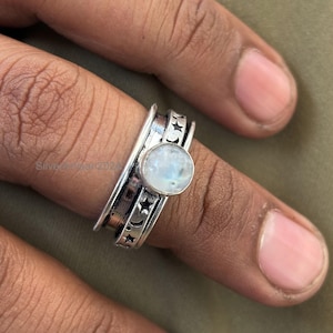 May include: A silver ring featuring a moonstone and a design of moons and stars. The ring is worn on a finger.
