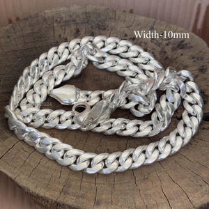 Puede incluir: Un collar de cadena de plata con un ancho de 10 mm. La cadena está formada por eslabones entrelazados y tiene un cierre de mosquetón. El texto "Width-10mm" es visible en la imagen.