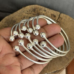 Op de afbeelding: Een verzameling zilverkleurige armbanden. Elke armband heeft een gebogen ontwerp met een kleine, ronde, zilveren kraal aan het einde van het gebogen gedeelte. De armbanden worden op een houten oppervlak gepresenteerd.