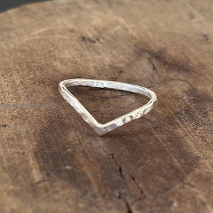 Może przedstawiać: Srebrny pierścionek w kształcie litery V z młotkowaną fakturą. Pierścionek jest umieszczony na tle ciemnego drewna. Pierścionek jest oznaczony znakiem "925".