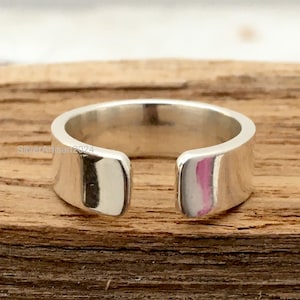 Könnte beinhalten: Ein silberner, offener Ring mit einem flachen, rechteckigen Band. Der Ring hat eine polierte, reflektierende Oberfläche. Der Ring wird auf einer Holzoberfläche präsentiert.