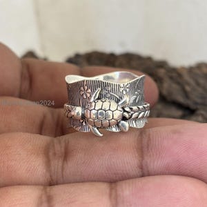 Könnte beinhalten: Ein silberner Drehring mit einem detaillierten Schildkrötenmotiv und floralen Akzenten. Der Ring wird an einem Finger getragen.
