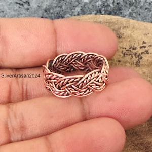 Puede incluir: Un anillo de cobre que presenta un diseño de metal trenzado con un acabado pulido. El anillo muestra un patrón continuo de hebras entrelazadas. El texto SilverArtisan2024 es visible en el lado izquierdo de la imagen cerca del anillo.