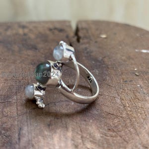 May include: A sterling silver ring featuring three gemstones. The ring has a unique design with a large, round, green-blue gemstone and two smaller, round, clear gemstones. The band is marked with "925".