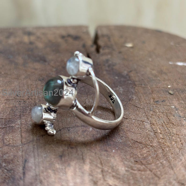 May include: A sterling silver ring featuring three gemstones. The ring has a unique design with a large, round, green-blue gemstone and two smaller, round, clear gemstones. The band is marked with "925".