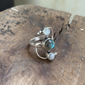 May include: A sterling silver ring featuring a central, oval-shaped labradorite stone with iridescent blue and green hues. The ring has a unique design with two smaller, round, white moonstone accents and the number "925" engraved on the band.