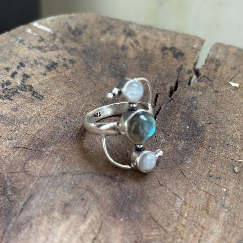 May include: A sterling silver ring featuring a central, oval-shaped labradorite stone with iridescent blue and green hues. The ring has a unique design with two smaller, round, white moonstone accents and the number "925" engraved on the band.