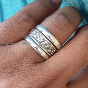 May include: A silver ring with a floral pattern. The ring has three bands, with the central band displaying an engraved design of flowers and leaves. The ring is on a finger, with the text "SilverArtisan2024" visible.