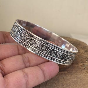 Könnte beinhalten: Ein silbernes Armreif-Armband mit detaillierten floralen und geometrischen Mustern. Das Armband hat geprägte Designs und eine polierte Oberfläche. Das dekorative Band ist etwa 1,9 cm breit und zeigt einen klassischen, eleganten Stil.