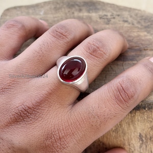 May include: A silver ring with a large, oval, deep red gemstone. The ring has a simple, polished band and a bezel setting that holds the stone securely. The gemstone is the focal point, catching the light and reflecting a rich color.