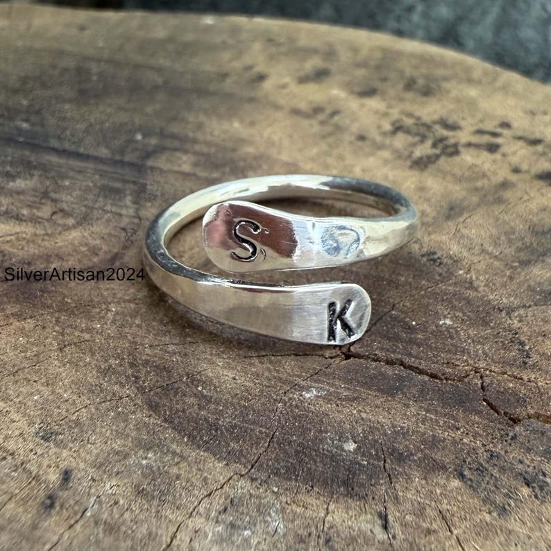 May include: A silver-colored bypass ring featuring a polished finish and two open ends. Each end is stamped with a single black letter, specifically the letter S on the top end and the letter K on the bottom end. The ring has a smooth, metallic surface.