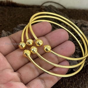 May include: A set of three gold-colored bangles. Each bangle has a smooth, rounded ball at the end of a curved, slender bar. The bangles are displayed on a hand, showcasing their bright, reflective surface. The jewelry is simple and elegant.