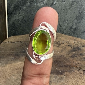 Può includere: Un anello in argento con una grande pietra preziosa color verde lime traslucido dal taglio ovale, incastonata in una fascia di metallo lucido con accenti curvi e organici. Il testo "SilverArtisan2024" è visibile a sinistra.