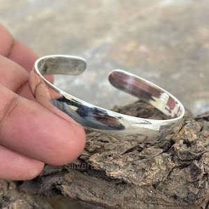 Puede incluir: Un brazalete de plata con un acabado liso y pulido. El brazalete está abierto y tiene una forma ligeramente curvada.