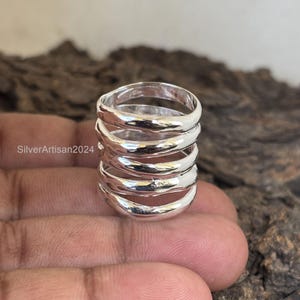 Könnte beinhalten: Ein silberner Ring mit einem breiten Band aus fünf übereinanderliegenden, gewellten und leicht unebenen Bändern.