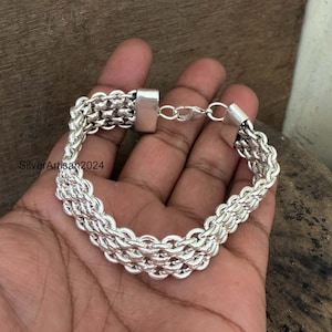 Peut inclure: Un bracelet à chaîne de couleur argentée présentant un design à maillons tissés à plusieurs brins. Le bracelet est composé d'anneaux métalliques circulaires interconnectés et comprend un fermoir en métal poli. Le texte SilverArtisan2024 est visible.