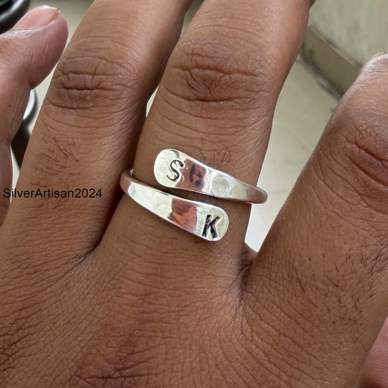 May include: A silver-colored bypass ring featuring two flat, rounded ends. Each end is stamped with a single black letter, with the letter S on the top end and the letter K on the bottom end. The ring has a smooth, polished metallic finish.