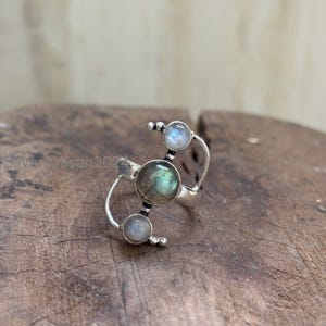 May include: A silver ring featuring a central, round, green-grey gemstone, flanked by two smaller, round, light-colored stones. The ring has a unique, open design with curved bands and small, beaded accents. The ring is set on a wooden surface.