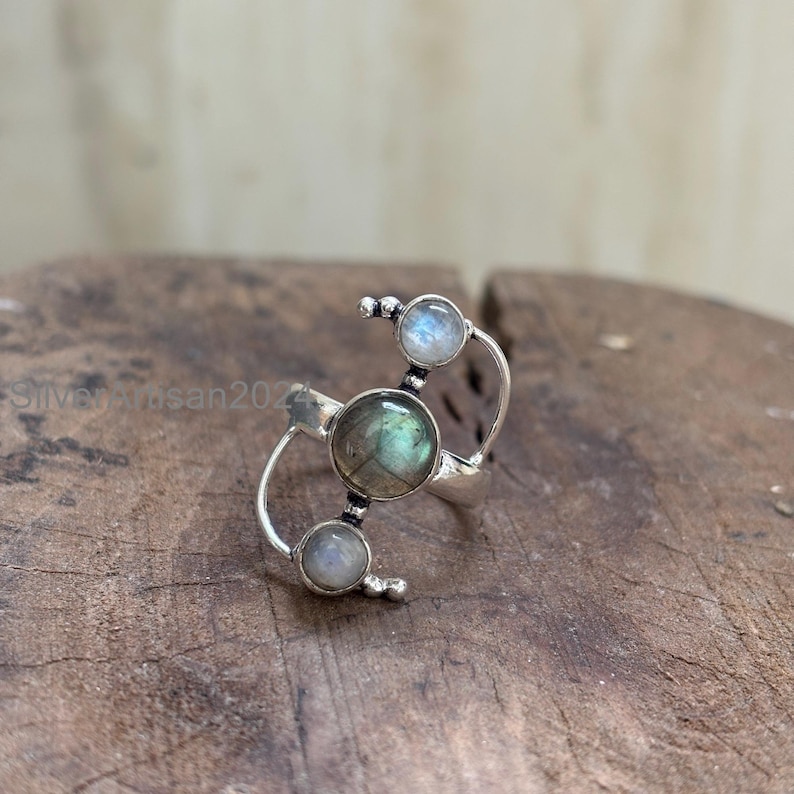 May include: A silver ring featuring a central, round, green-grey gemstone, flanked by two smaller, round, light-colored stones. The ring has a unique, open design with curved bands and small, beaded accents. The ring is set on a wooden surface.