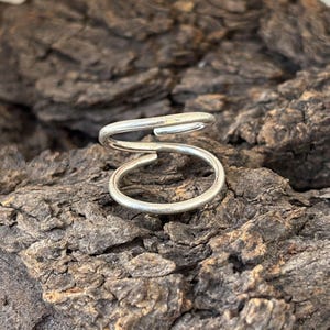 May include: A silver double-loop ring, made of twisted wire, is shown on a rough wooden surface.
