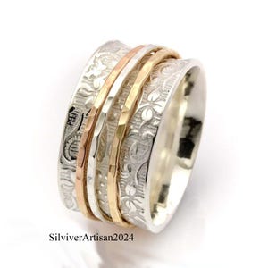 May include: A silver spinner ring with floral engravings and multiple bands in gold and rose gold. The ring has a textured design and a polished finish. The text "SilviverArtisan2024" is visible on the ring.