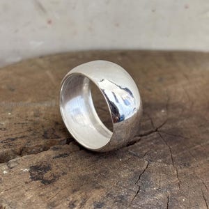 May include: A polished, wide silver ring with a rounded top. The ring reflects light, creating highlights on its surface. The ring is set against a rustic, wooden background, showcasing its simple elegance.