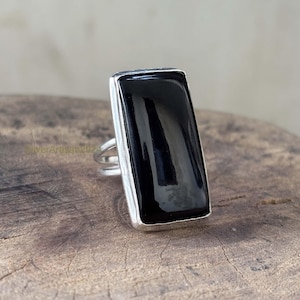 May include: A rectangular black onyx ring with a silver band. The polished black stone is set in a silver bezel, creating a striking contrast. The ring is displayed on a wooden surface.