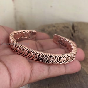 Puede incluir: Un brazalete de color cobre con un diseño tejido. El brazalete abierto está hecho de piezas de metal curvadas e entrelazadas. El brazalete se sostiene en una mano.