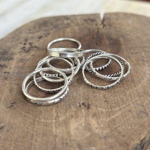 May include: A collection of silver-toned metal rings featuring various designs, including smooth bands, twisted rope textures, and patterned surfaces. The rings are stacked and overlapping, showcasing different widths and intricate detailing.