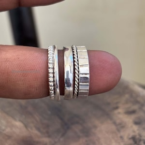 Może przedstawiać: Kolekcja srebrnych pierścionków o różnych fakturach i wzorach. Pierścionki mają wzory koralikowe, gładkie, skręcone i prostokątne. Pierścionki są prezentowane na palcu.